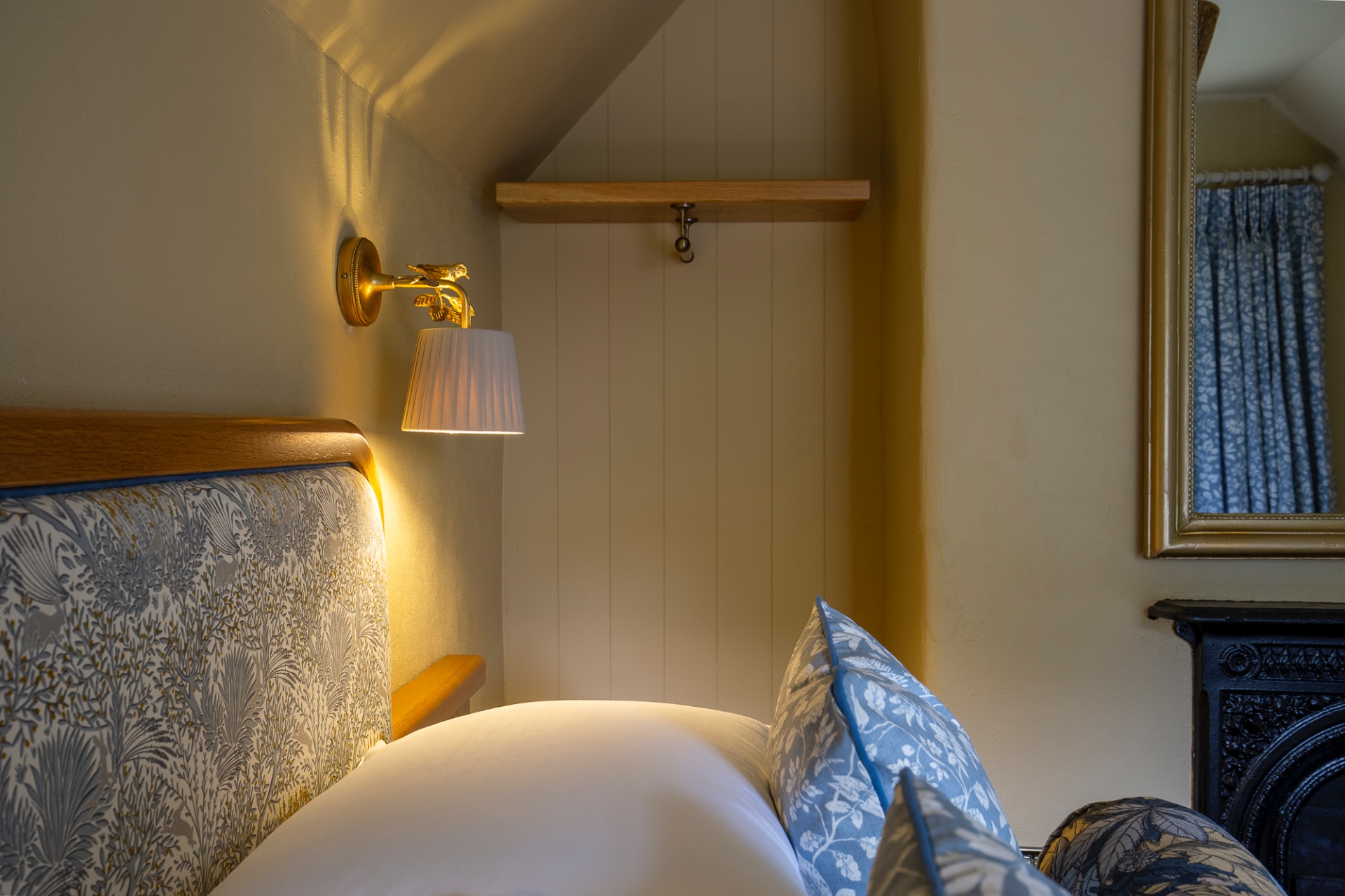 Warm-lit bedroom corner with brass wall lamp, patterned headboard, and blue curtains beside a mirror.