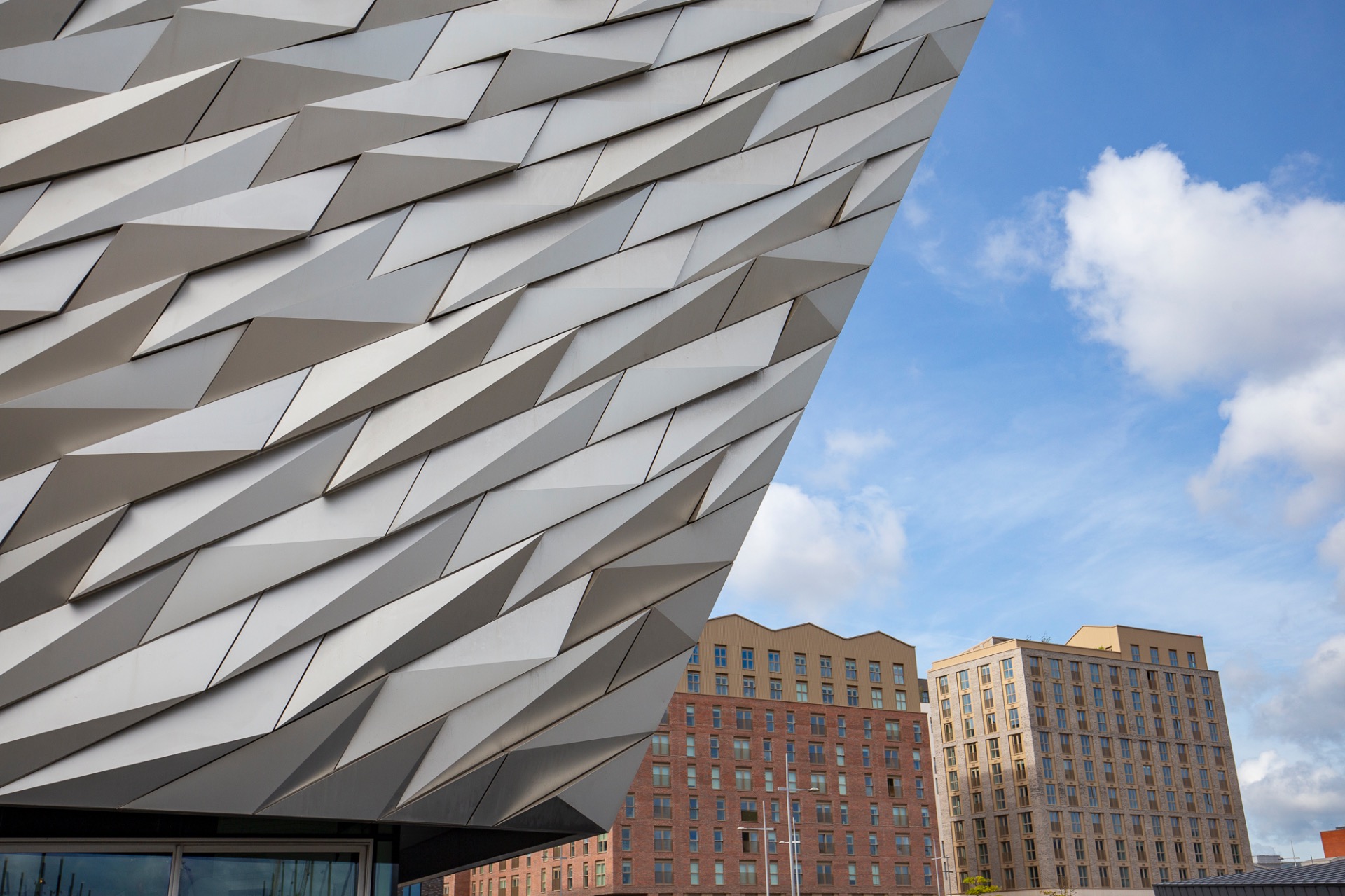 Geometric gray metal facade with angular panels, with brick and glass buildings and blue sky behind.